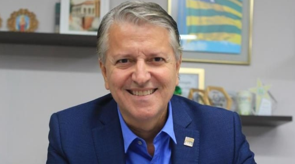 Retrato de Joel de Sant'anna Braga Filho, secretário de Indústria e Comércio de Goiás, sorrindo em um escritório com uma bandeira de Goiás ao fundo.